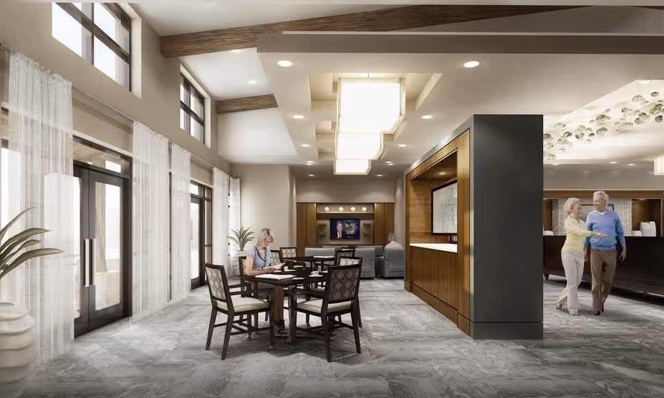 Spacious interior common area of a senior living facility with tables, chairs, lounge seating, and residents near large windows.