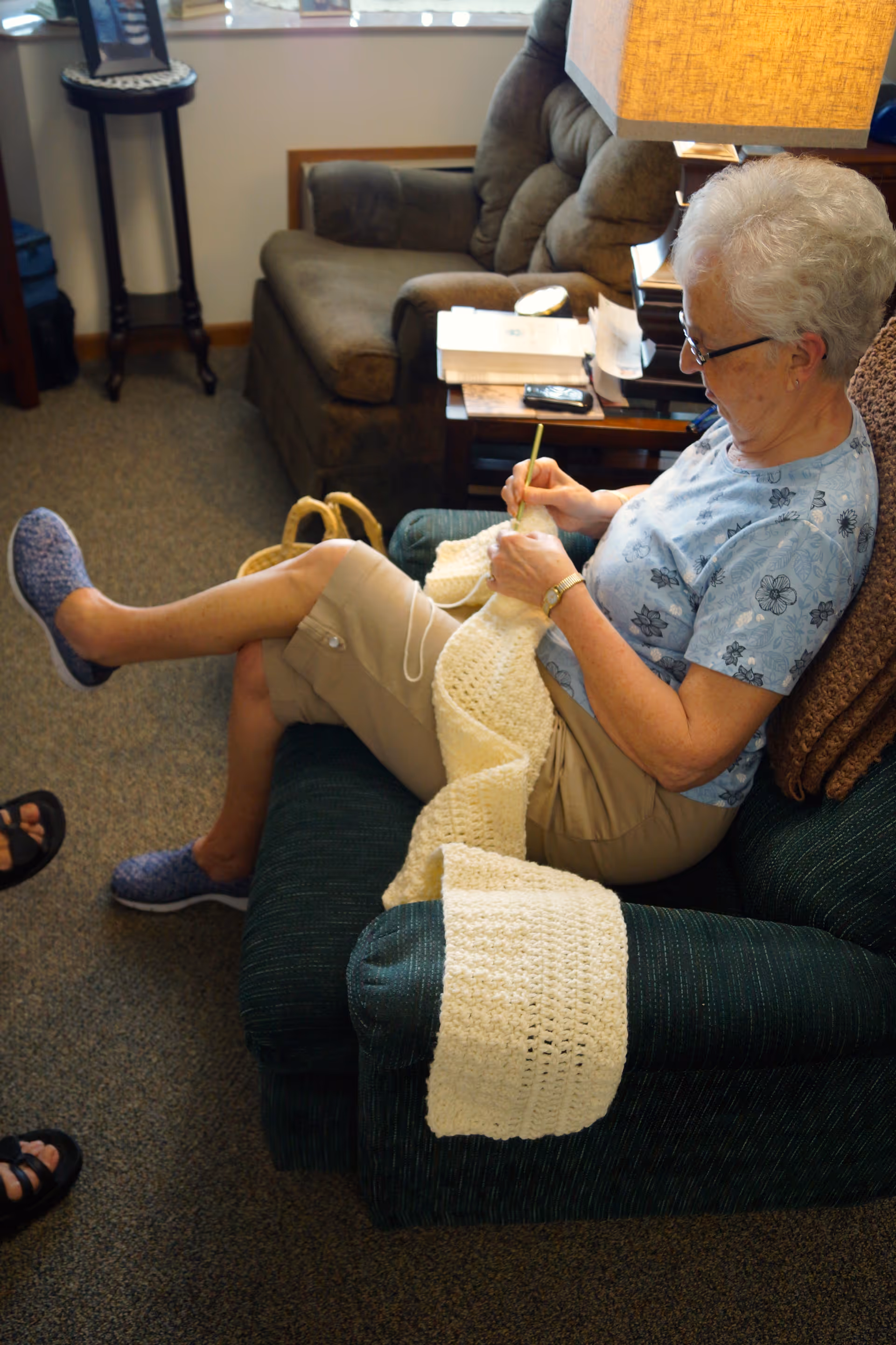 An elderly woman with short white hair and glasses is sitting on a green armchair in a cozy living room, crocheting a large cream-colored blanket. She is wearing a light blue floral shirt, beige shorts, and blue slippers. Behind her is a brown armchair, a side table with books, a lamp, and a window with blinds partially open.