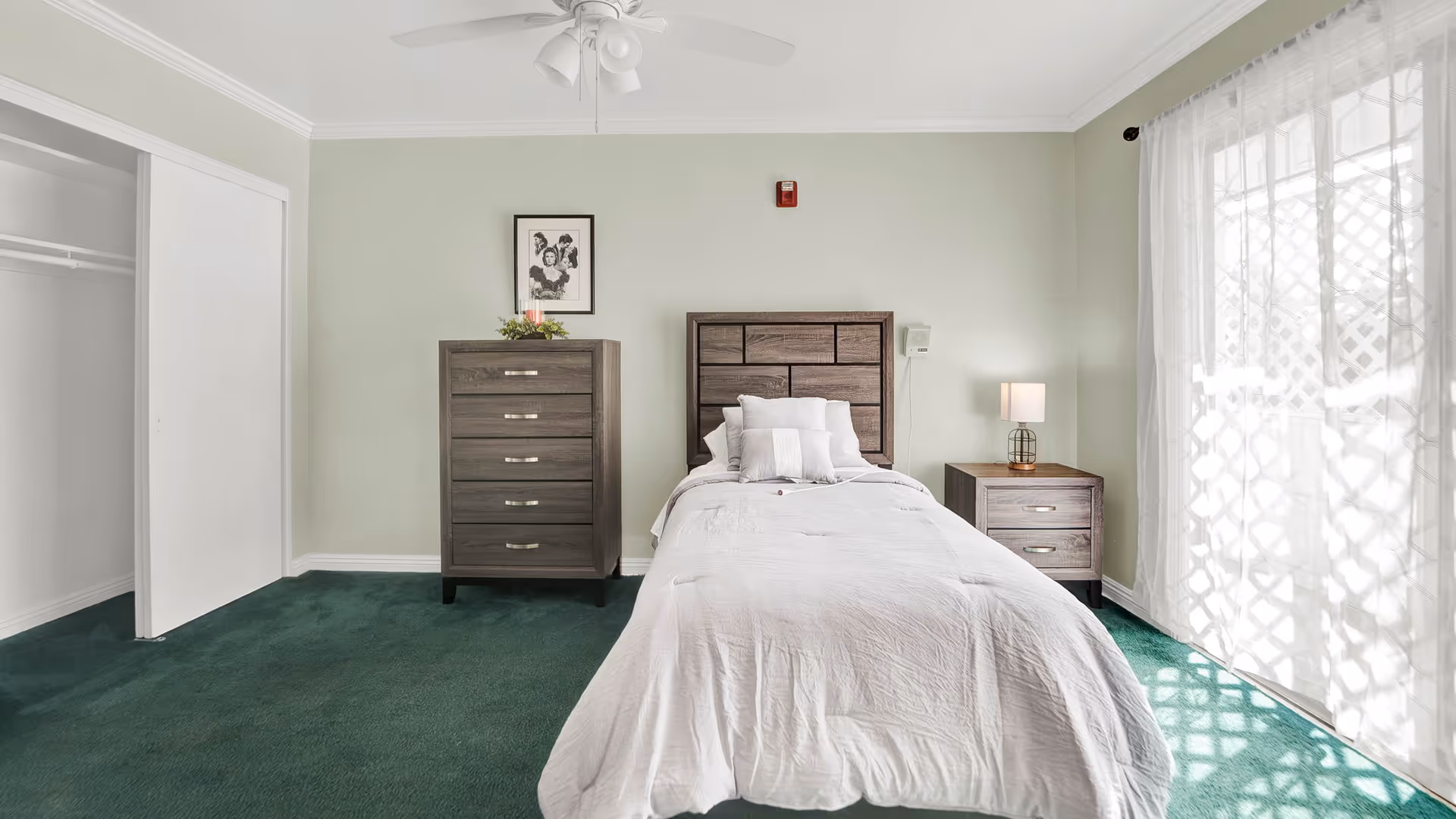 A bright bedroom with a single bed, wooden headboard, matching dresser and nightstand, green carpet, and a sliding glass door with sheer curtains.