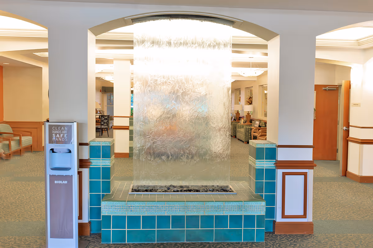 Interior view of a senior living facility lobby area featuring a decorative water wall with textured glass and turquoise tiled base. To the left, there is a hand sanitizer dispenser with a sign that reads 'Clean Hands Are Safe Hands.' The background shows seating areas with chairs and tables, warm lighting, and wooden accents on the walls and door frames.