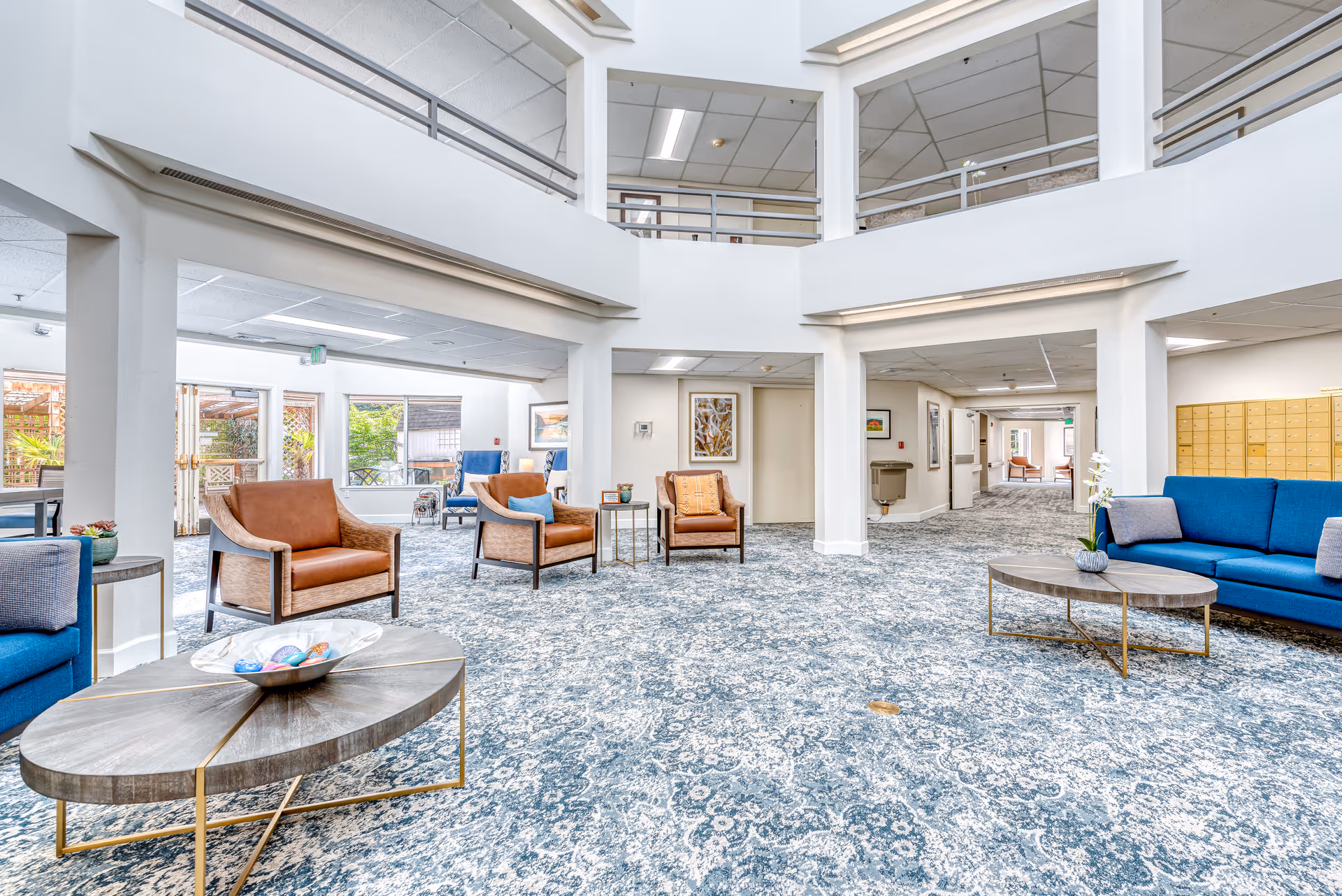 A spacious and bright senior living facility common area with patterned blue carpet, white walls, and a high ceiling with an open second-floor balcony. The room is furnished with blue and brown armchairs, two oval wooden coffee tables with decorative items, and artwork on the walls. Large windows and glass doors allow natural light to enter, and there is a hallway leading to other parts of the facility.