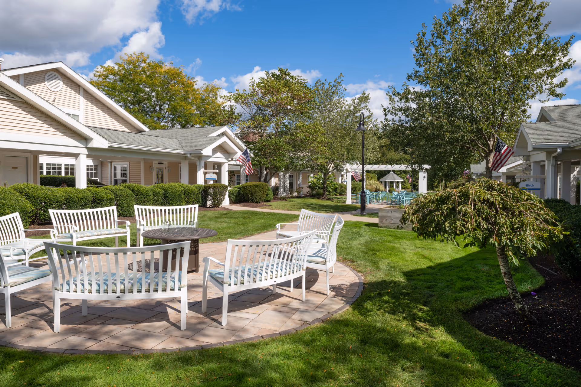 A sunny outdoor courtyard with white patio benches arranged around a circular paver area, surrounded by single-story cottage buildings and lawns.