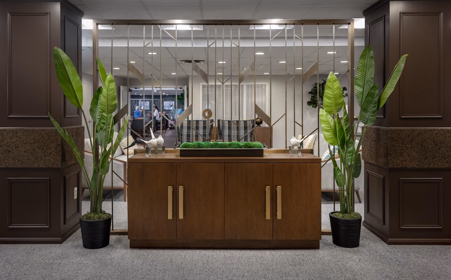 Interior view of a senior living community lounge area with a wooden cabinet topped with decorative greenery and two potted plants on either side. Behind the cabinet is a decorative metal divider, and in the background, there are seating areas with chairs and people engaged in conversation.