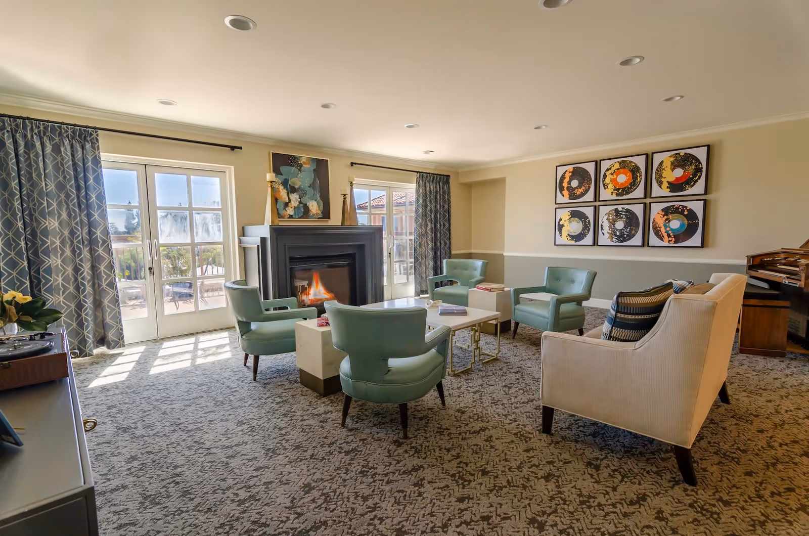 A cozy living room with a fireplace, surrounded by four light green armchairs and a beige sofa. The room has patterned carpet flooring, large windows with blue patterned curtains, and modern artwork on the walls. There is a piano in the corner and a coffee table in the center with books on it.