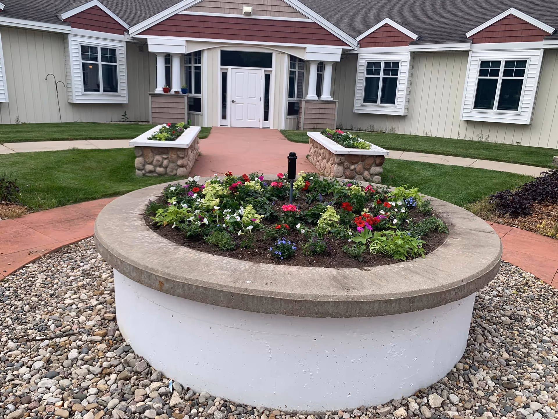 Outdoor view of a senior living facility entrance with a circular flower bed filled with colorful flowers in the foreground, surrounded by a stone border and pebbles. The building has beige siding with white trim and red accents, multiple windows, and a white double door entrance with columns.