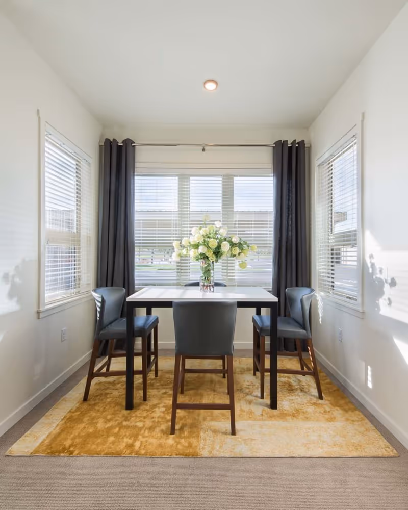 A bright dining area with a square white table surrounded by four dark gray chairs. The table has a vase with white flowers in the center. The room has three windows with white blinds and dark curtains on the middle window. A yellow and beige rug is underneath the table and chairs.