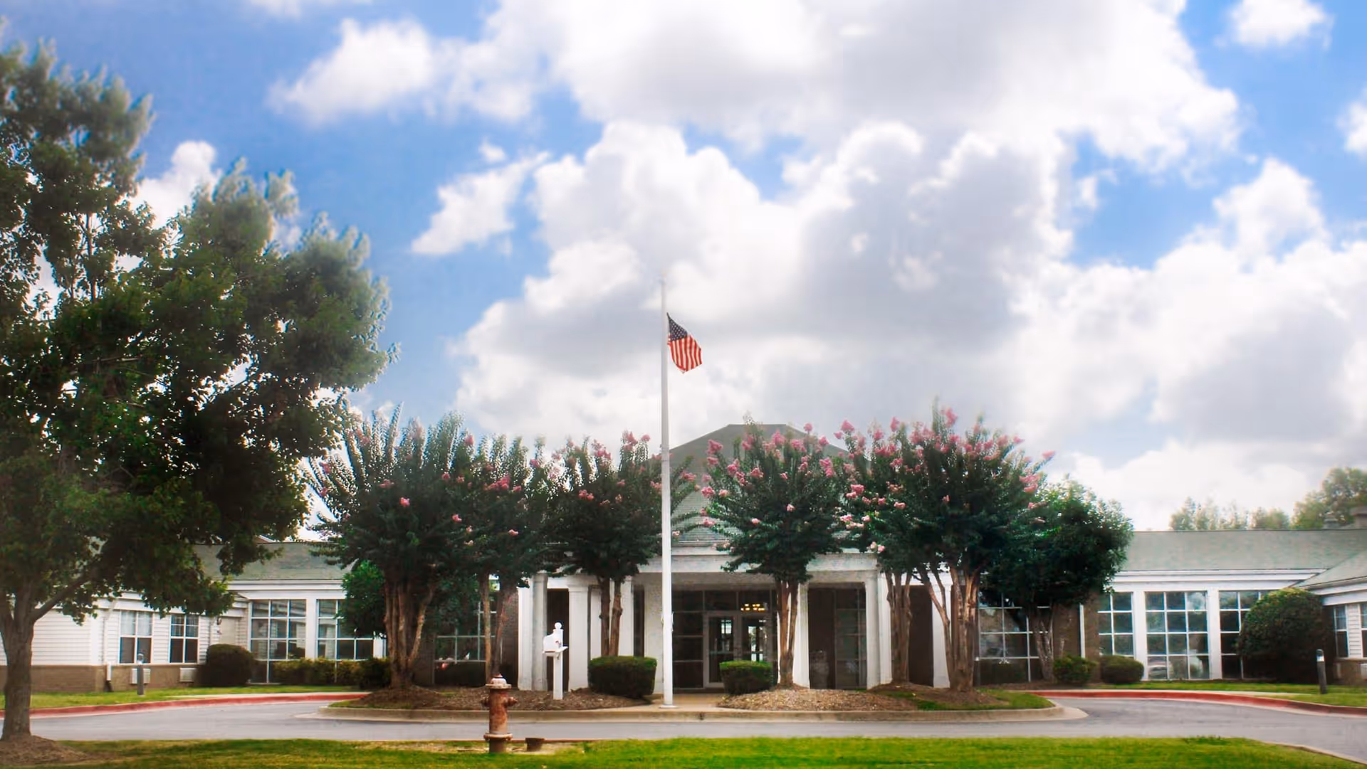 Front entrance of a single-story senior living facility with a flagpole, flowering trees, and a circular driveway under a partly cloudy sky.
