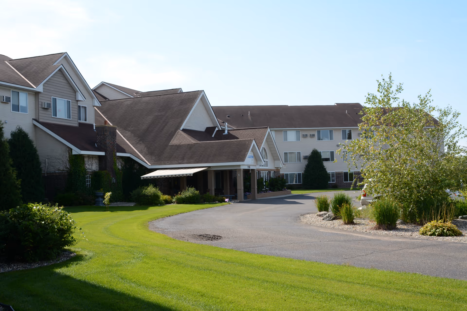 Exterior front of a multi-story senior living facility with a covered entrance, curved driveway, and landscaped lawn.