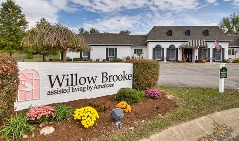 Exterior view of Willow Brooke assisted living facility by Americare, showing a white building with black shutters, an American flag, and a landscaped area with colorful flowers and a sign displaying the facility's name.