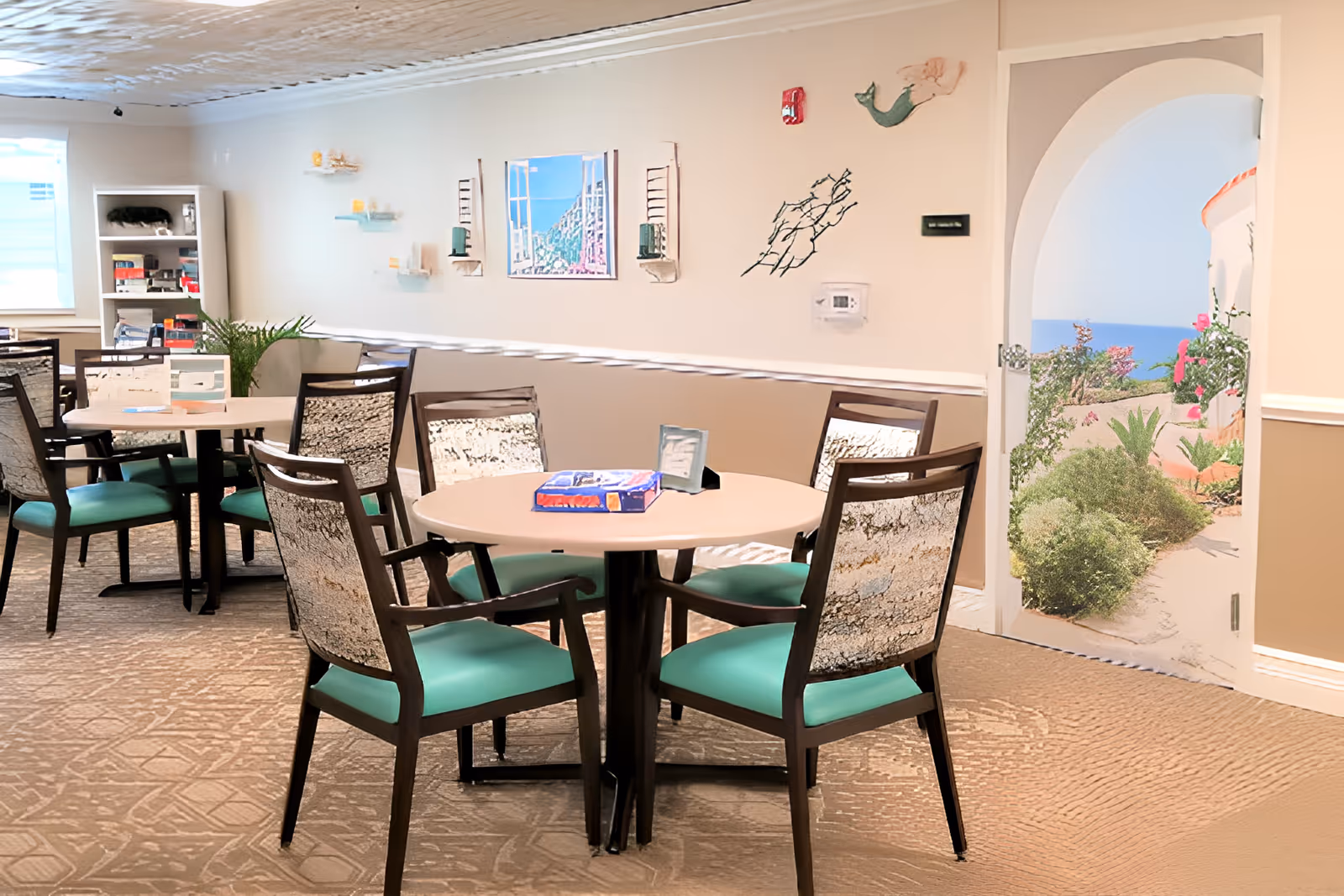 A dining room with round tables and chairs featuring green cushions and patterned backs. The room has beige walls decorated with small shelves, artwork, and a wall-mounted thermostat. There is a bookshelf in the corner and a door with an arched frame that opens to a garden path with greenery and a view of the ocean.
