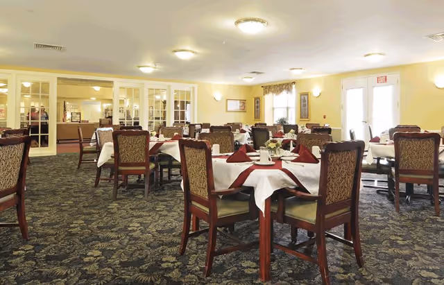 A spacious dining room with multiple tables covered in white tablecloths and burgundy napkins. Each table is surrounded by wooden chairs with patterned upholstery. The room has yellow walls, carpeted floors with a floral pattern, and large windows letting in natural light. There are ceiling lights and wall sconces providing additional lighting.