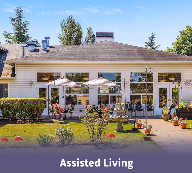 Exterior view of a single-story assisted living facility building with large windows, outdoor seating with tables and umbrellas, a multi-tiered fountain, potted plants, and lawn decorations including pink flamingos under a clear blue sky.