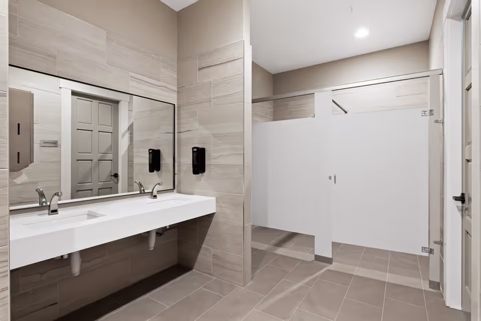 Modern public restroom with two sinks and faucets mounted on a white countertop, large mirror above the sinks, beige tiled walls and floor, two soap dispensers on the wall, and two toilet stalls with white doors and partitions.