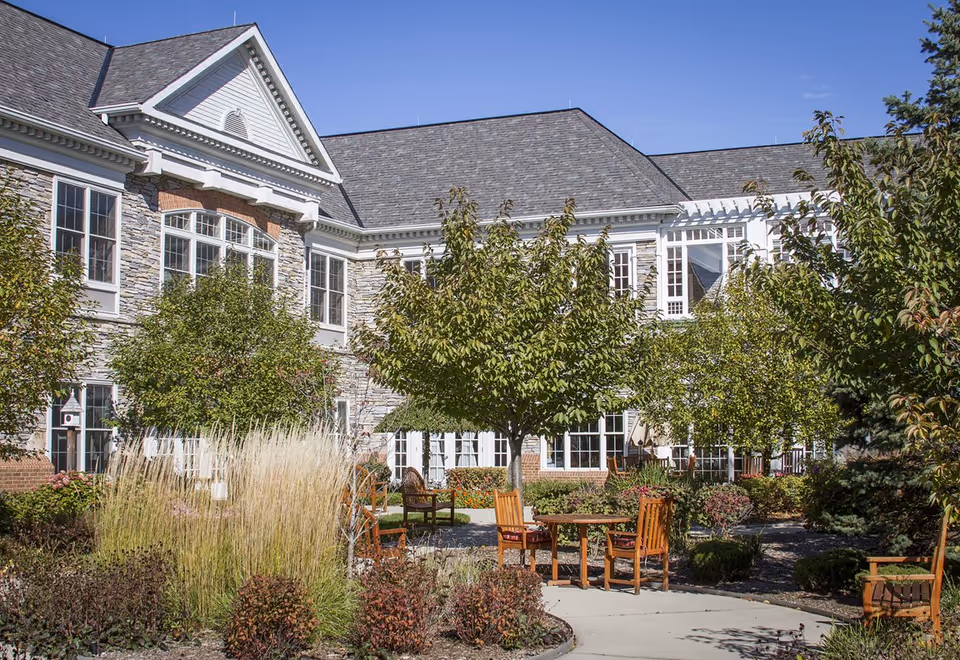 Outdoor garden area at Sunrise of Grosse Pointe Woods featuring a paved walkway, wooden chairs and tables, various trees, shrubs, and ornamental grasses, with a large stone and brick building in the background under a clear blue sky.