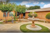 Exterior courtyard with a central stone fountain, circular grass islands, trimmed trees, benches and a low brick building.