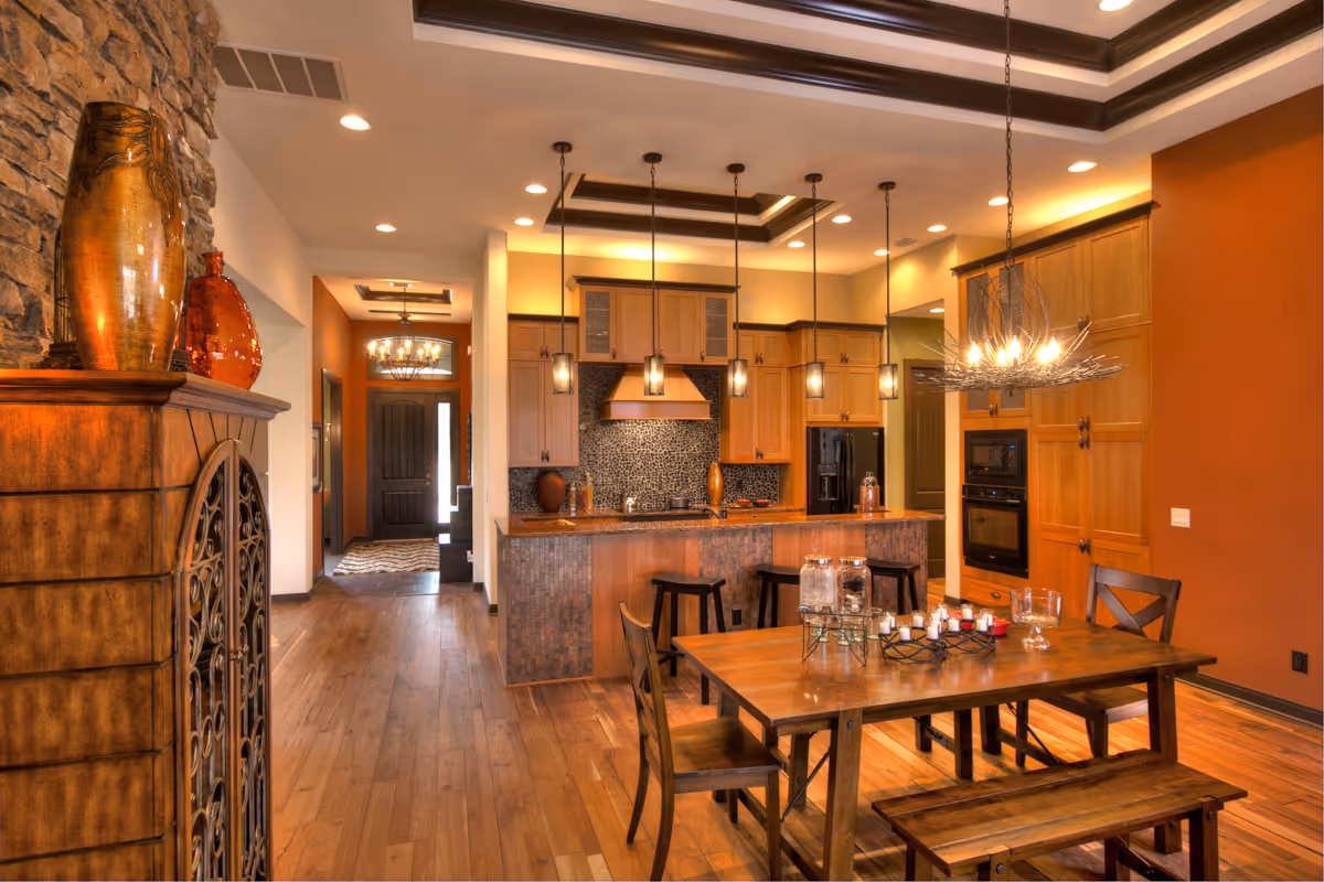 A warm, inviting kitchen and dining area with wooden floors and cabinetry. The kitchen features a central island with bar stools and pendant lighting above. The dining area has a wooden table with chairs and a bench, decorated with candles and glass jars. The walls are painted in earthy tones, and there is a stone fireplace with decorative vases on the left side.
