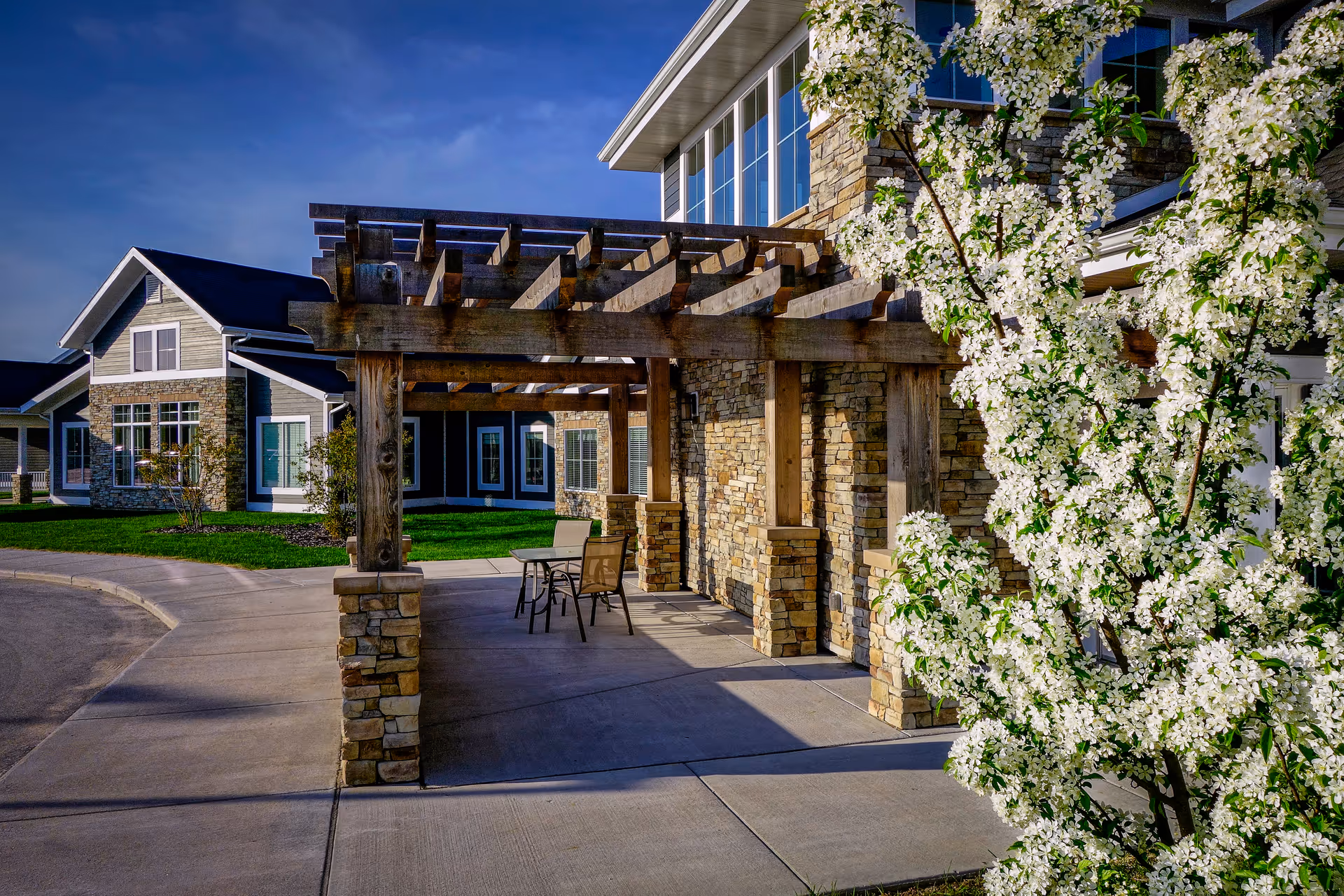 Outdoor view of Sheboygan Senior Community showing a stone and wood pergola with a table and chairs underneath, a blooming white-flowered tree on the right, and residential-style buildings with large windows and stone accents in the background under a clear blue sky.