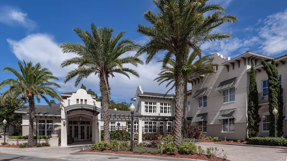 Exterior view of a senior living facility named The Estate at Hyde Park, featuring a light-colored building with multiple windows, palm trees, and a clear blue sky with some clouds.