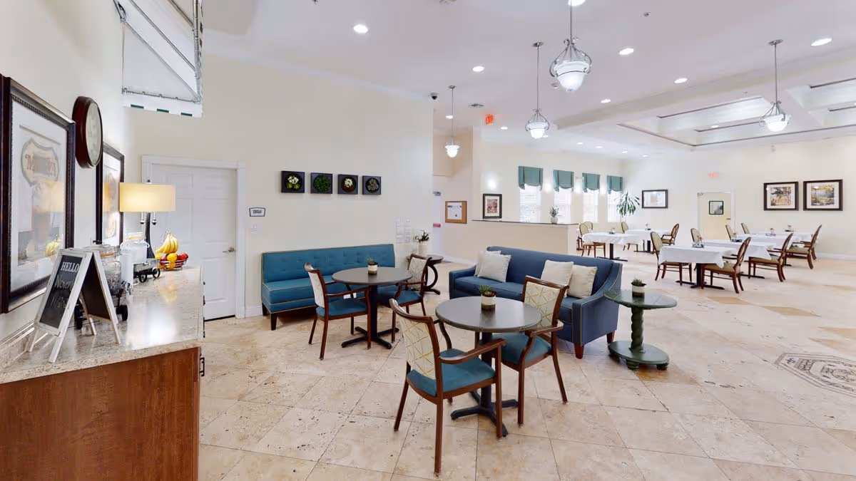Bright open dining and lounge area with round tables, upholstered chairs and sofas, and a buffet counter.