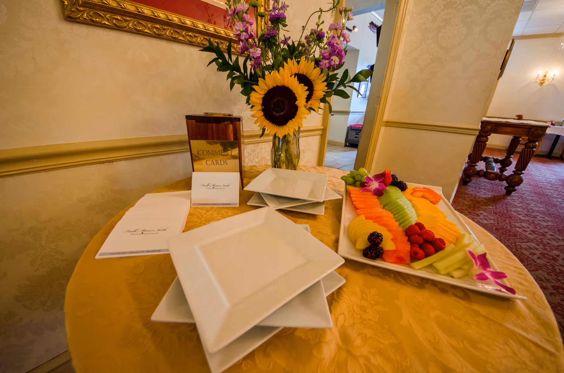 A round table covered with a yellow tablecloth displaying a platter of assorted sliced fruits including watermelon, cantaloupe, honeydew, pineapple, grapes, blackberries, and raspberries, decorated with purple orchids. There are several stacked white square plates, a vase with sunflowers and other flowers, and a wooden holder with comment cards on the table. The setting is indoors with a patterned carpet and cream-colored walls with molding.