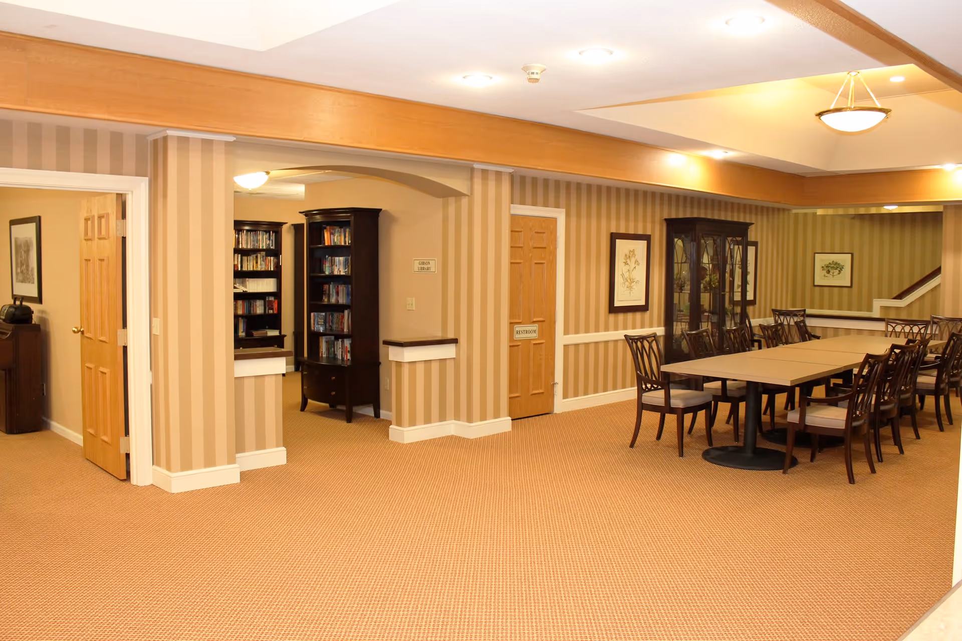 A spacious interior room with beige striped wallpaper and carpeted floor. There is a long table with multiple chairs arranged around it, a wooden cabinet with glass doors, and framed artwork on the walls. Two wooden doors are visible, one labeled 'RESTROOM' and the other 'LIBRARY'. Bookshelves filled with books are seen through an open archway.
