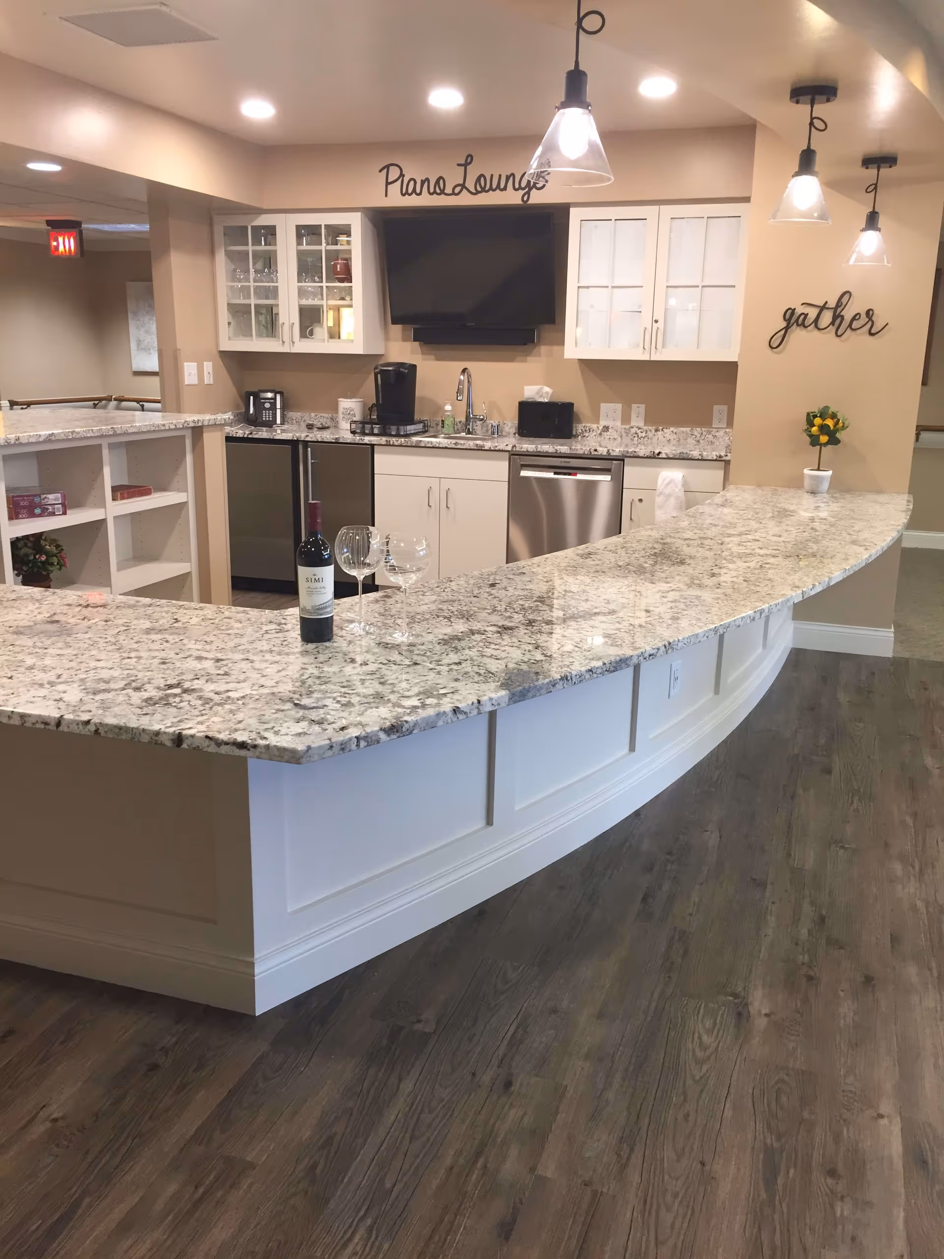 Interior view of a modern kitchen lounge area with a curved granite countertop bar, three hanging pendant lights, white cabinets with glass doors, a mounted flat-screen TV, a coffee maker, a dishwasher, and a bottle of wine with two wine glasses on the counter. The walls have decorative words 'Piano Lounge' and 'gather'.