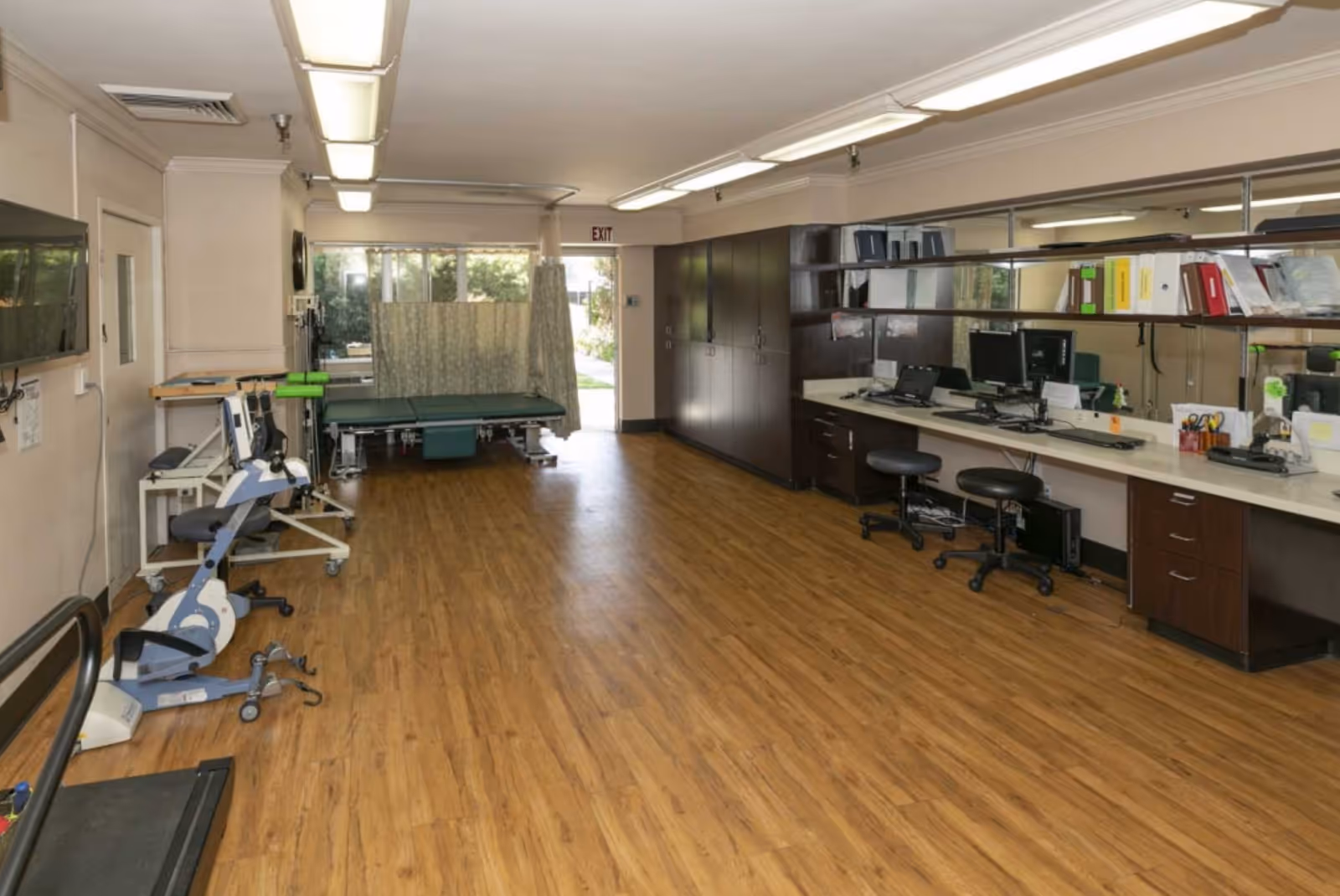 Interior view of a rehabilitation center room with wooden flooring, exercise equipment including a stationary bike and treadmill on the left, a treatment table with a privacy curtain near the back, and a long counter with computer workstations and office supplies on the right side.