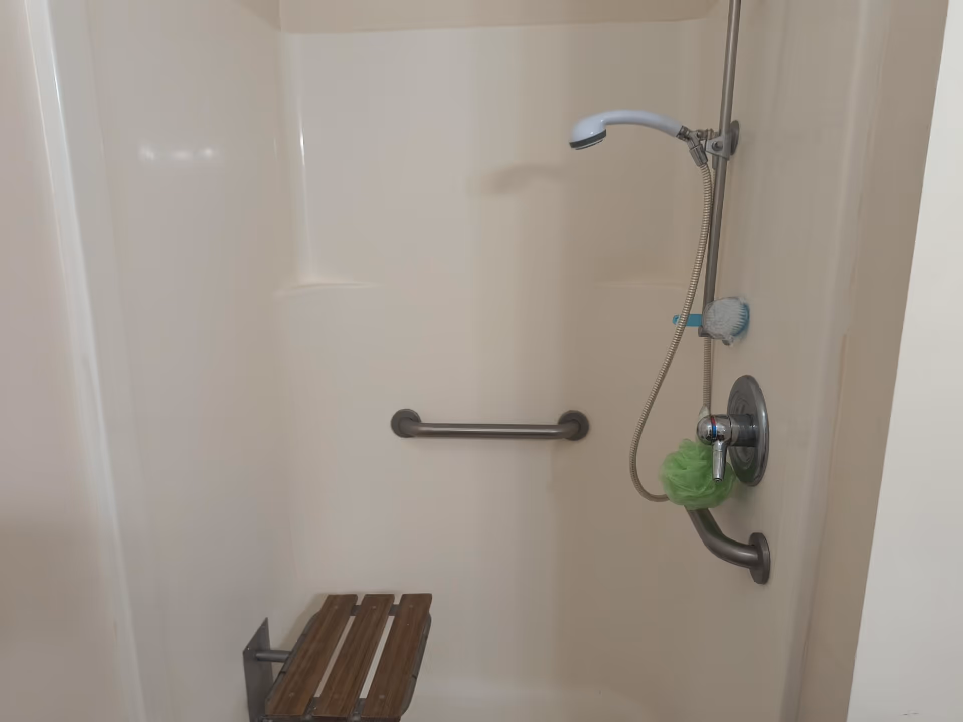 A shower area with white walls, a handheld showerhead attached to a vertical metal bar, a green bath pouf hanging on the faucet, a blue brush on the wall, a horizontal grab bar, and a foldable wooden shower seat.