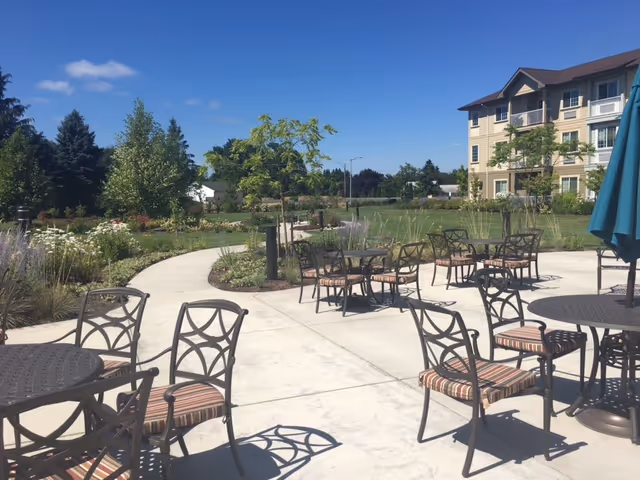 Outdoor patio area with metal tables and chairs featuring striped cushions, set along a curved concrete pathway surrounded by landscaped greenery and trees. A multi-story building is visible in the background under a clear blue sky.