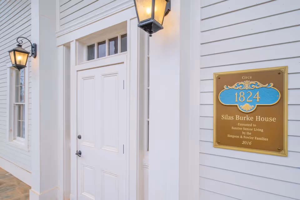 White exterior entrance with a closed door, two lit lanterns, and a plaque reading "Silas Burke House 1824" on the siding.