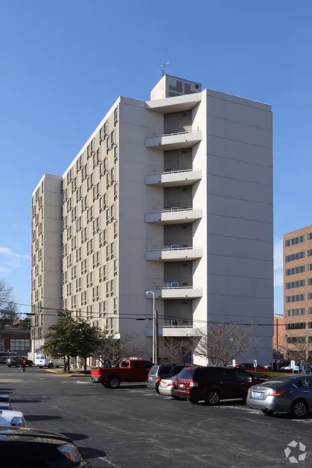Exterior view of a multi-story residential building with balconies on one side, surrounded by a parking lot with several parked cars under a clear blue sky.