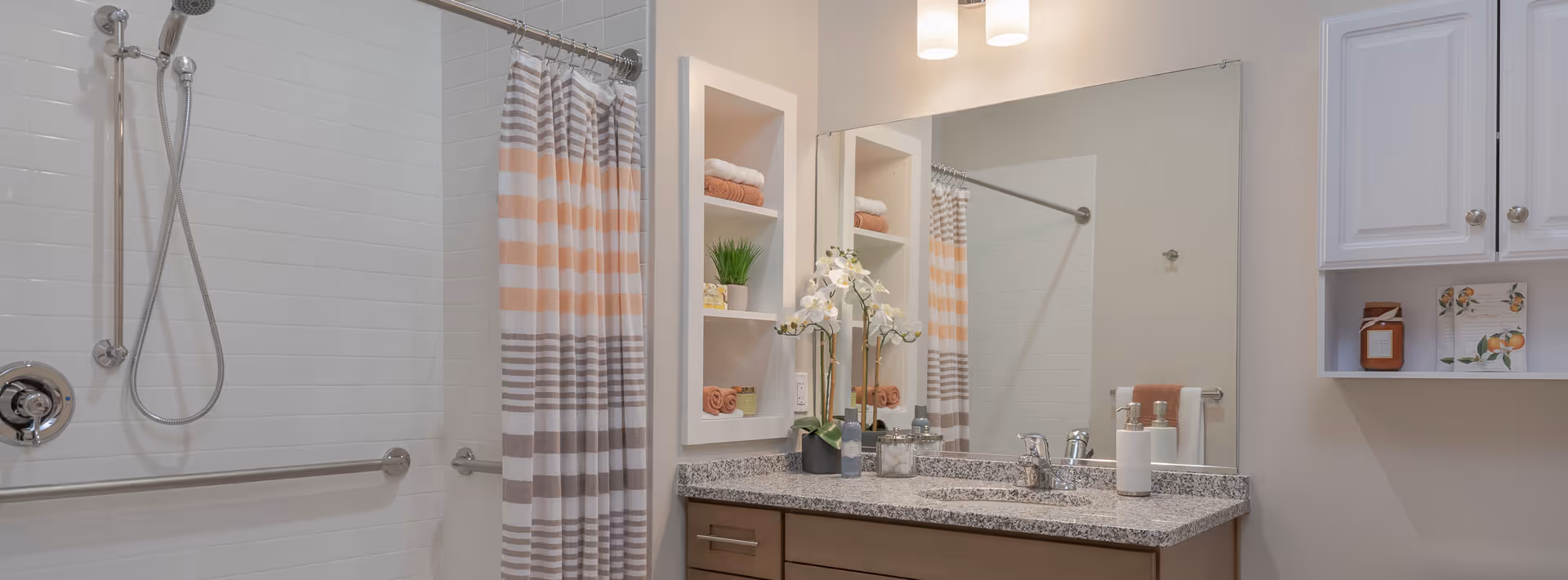 A clean and modern bathroom featuring a shower with a striped shower curtain in shades of gray, white, and peach. There is a granite countertop with a sink, soap dispensers, and a decorative orchid plant. Above the sink is a large mirror with two wall-mounted lights. Built-in shelves next to the mirror hold neatly folded towels and a small plant. White cabinets are mounted on the wall to the right.
