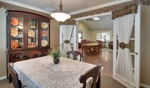 Dining room with a wooden table covered by a white lace tablecloth and four chairs. A small plant centerpiece is on the table. To the left is a wooden china cabinet displaying plates and glassware. The room opens through double doors with white curtains tied back, leading to a larger common area with additional tables and chairs.