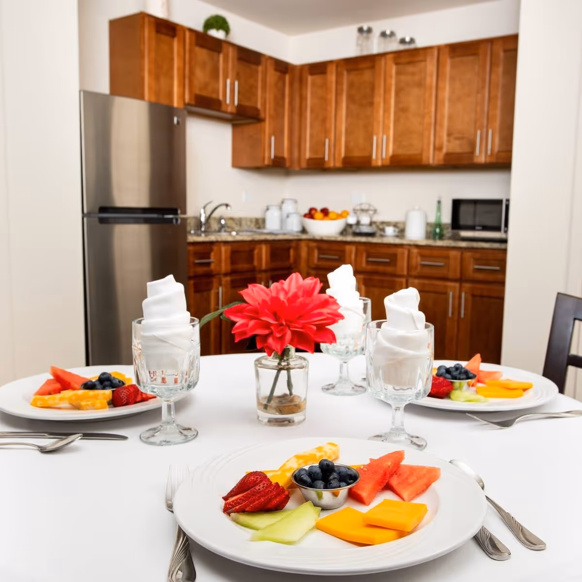 A dining table set with plates of assorted fresh fruit including strawberries, blueberries, watermelon, cantaloupe, and cheese slices. Each place setting has a glass with a neatly folded white napkin. In the center of the table is a small vase with a bright red flower. In the background, there is a kitchen with wooden cabinets, a stainless steel refrigerator, a microwave, and a bowl of fruit on the countertop.