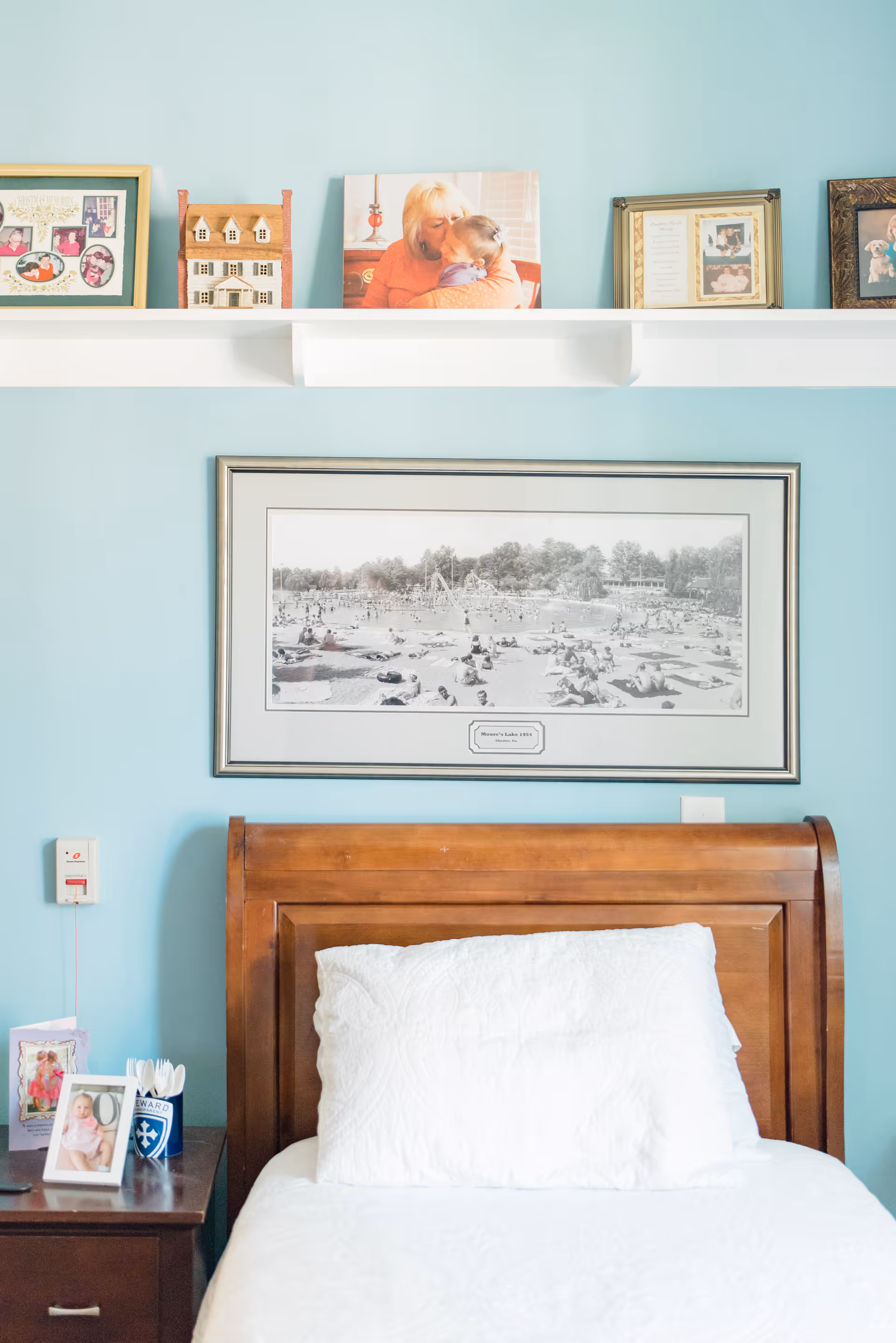 A neatly made single bed with a wooden headboard against a light blue wall. Above the bed is a large framed black and white photo of a beach scene with many people. A white shelf above the photo holds several framed pictures and a small wooden house model. A nightstand next to the bed holds framed photos and a cup with utensils.