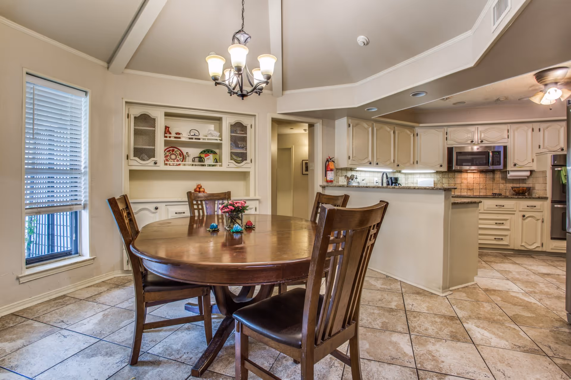 A dining area with a round wooden table and four matching chairs on a tiled floor. A chandelier with five lights hangs above the table. Behind the table is a built-in cabinet with decorative plates and other items. To the right is a kitchen with cream-colored cabinets, a microwave, and a tiled backsplash.