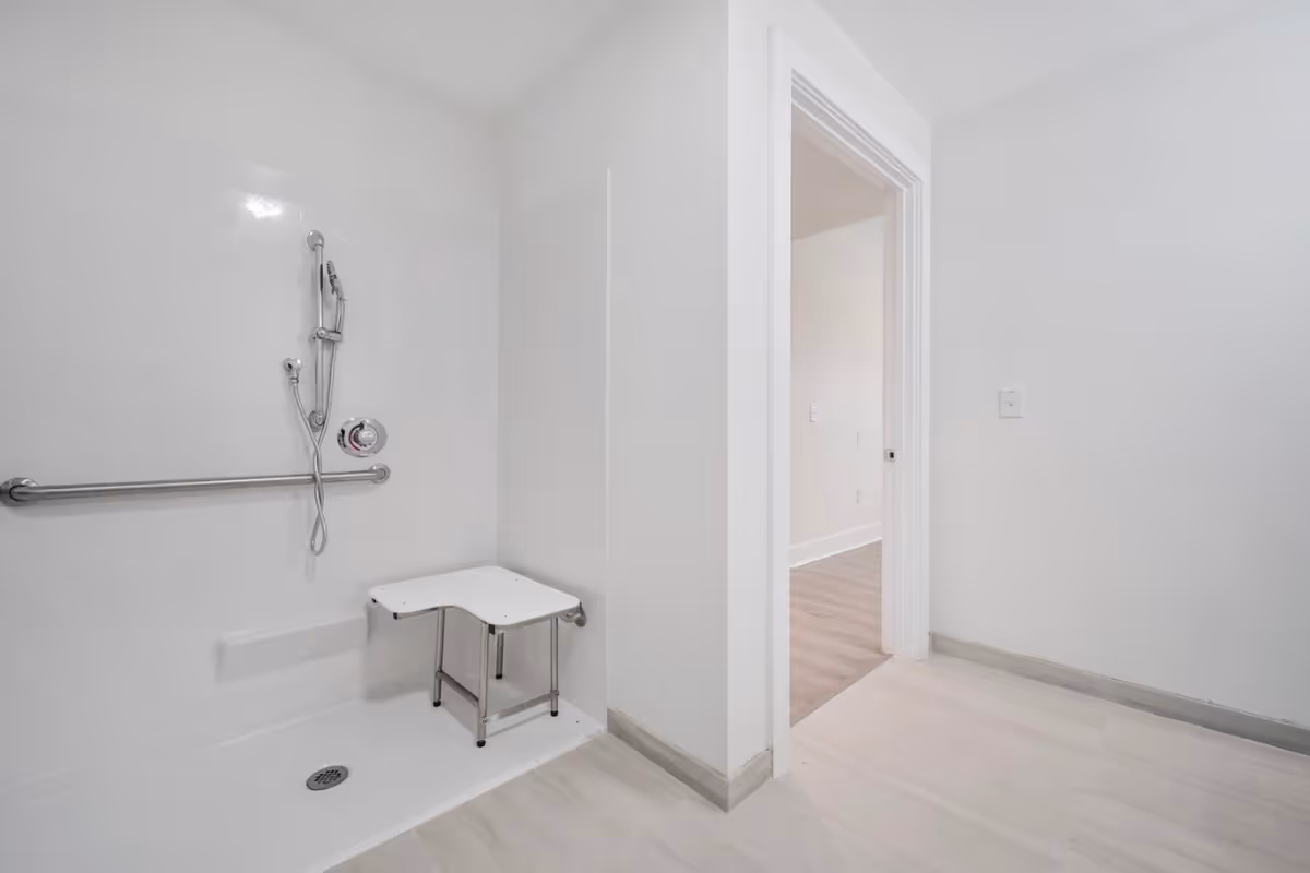 Wheelchair-accessible bathroom with a roll-in shower, grab bar, handheld shower head and a shower seat, viewed toward an open doorway.