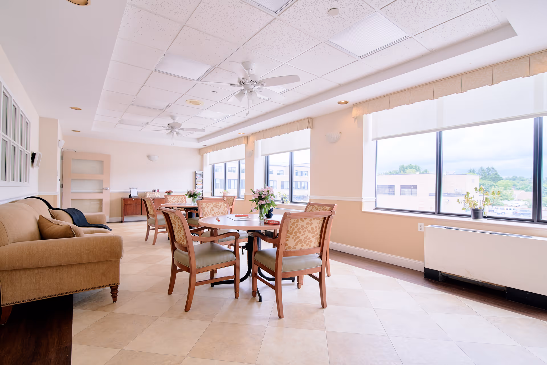 A bright and spacious common area with large windows letting in natural light. The room features several round tables with cushioned chairs arranged around them, a beige sofa on the left, ceiling fans, and light-colored tiled flooring. There are plants on the tables and a cabinet against the far wall.