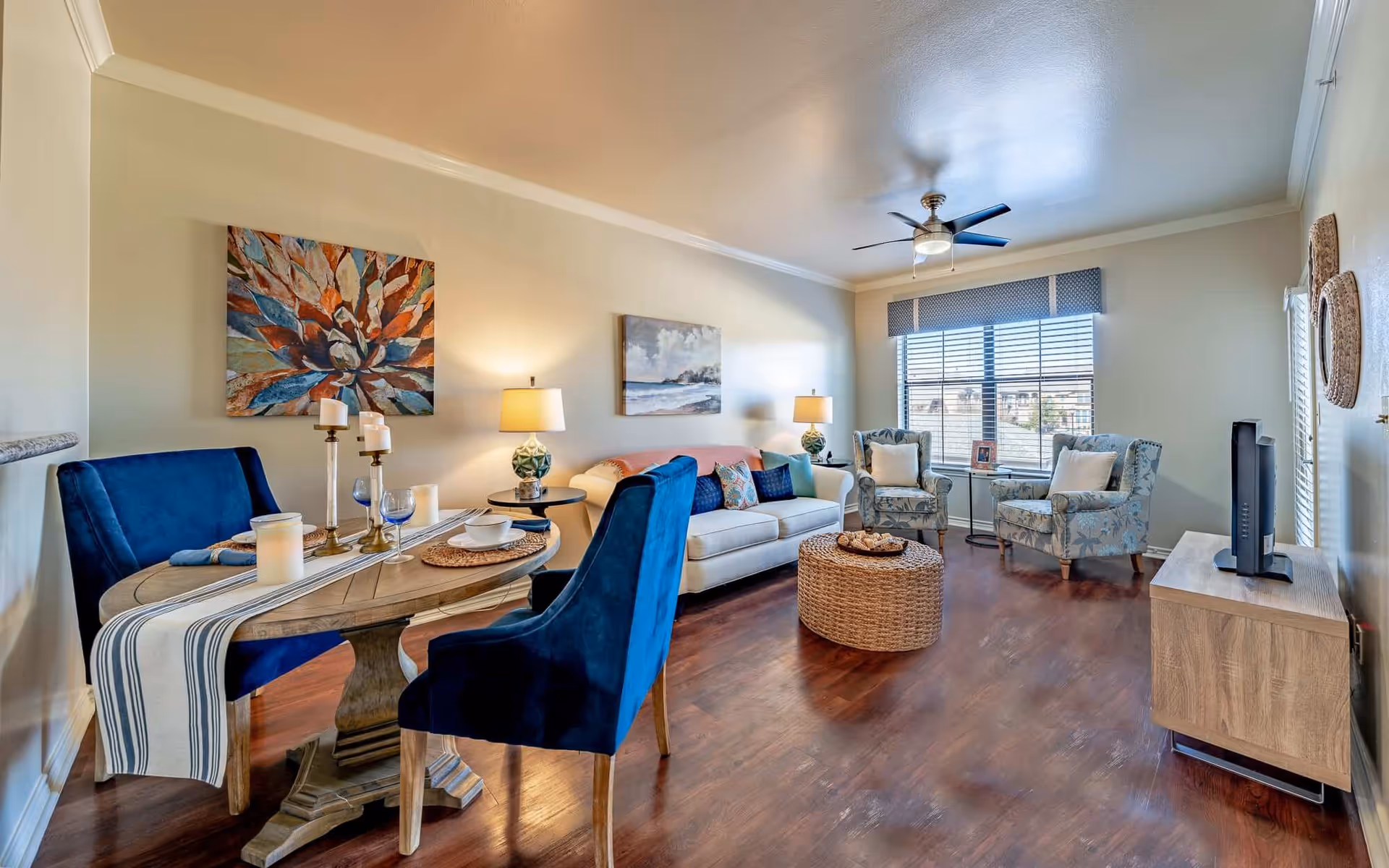 Sunlit living room and dining area with blue upholstered chairs, a white sofa, patterned armchairs, a round woven coffee table, and a TV cabinet.