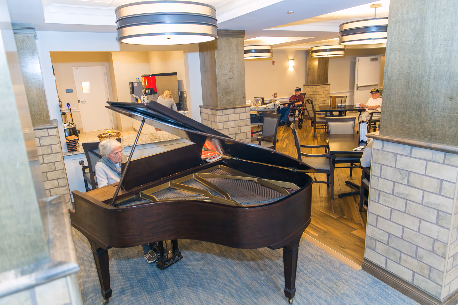 An elderly woman playing a grand piano in a common area of a senior living facility. The room has modern lighting fixtures, brick pillars, and several tables and chairs where other elderly residents are seated. A counter with a coffee machine is visible in the background.