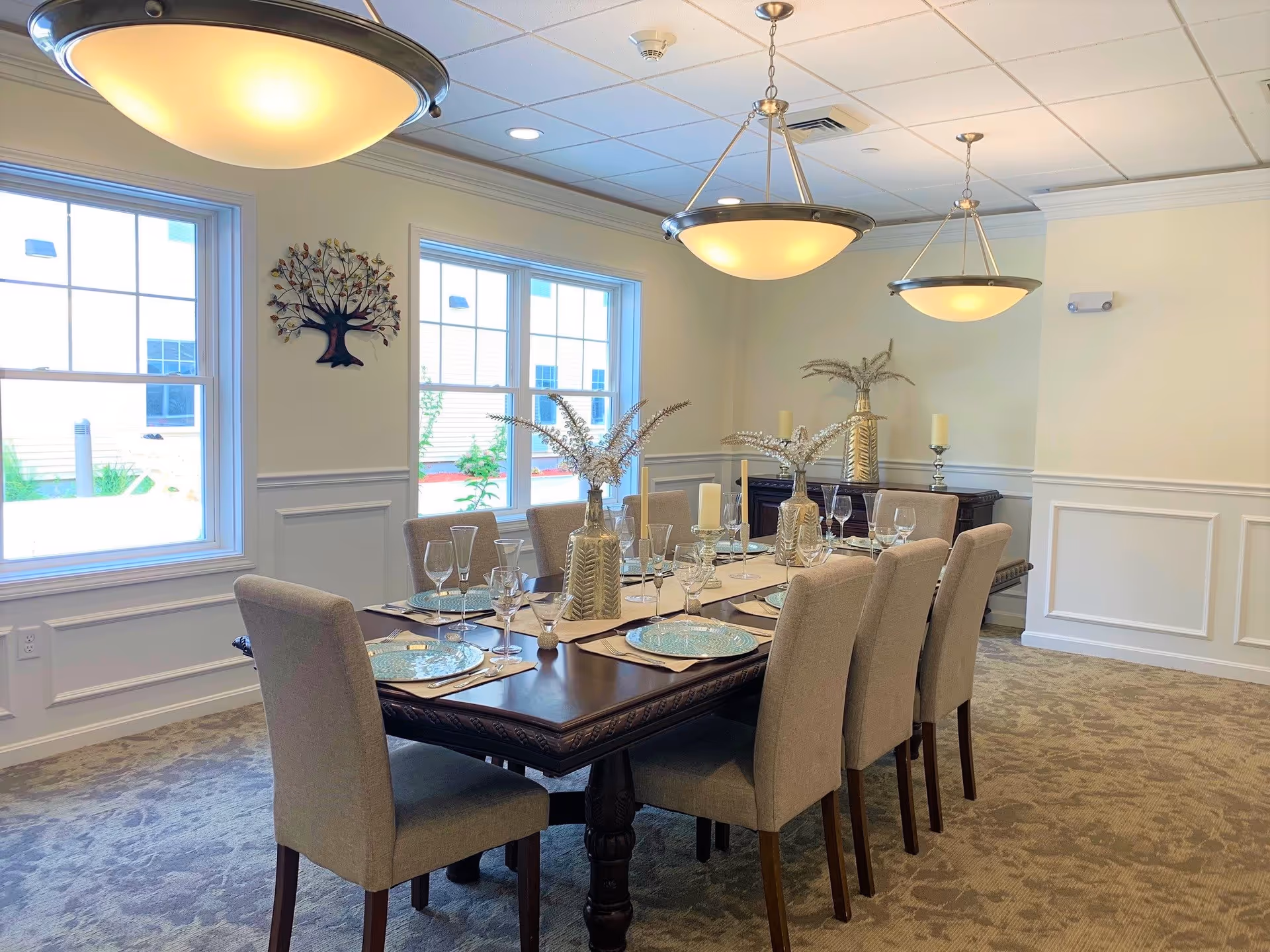 A well-lit dining room with a long wooden table set for eight people. The table is decorated with beige placemats, blue patterned plates, various glassware, candles, and vases with dried floral arrangements. The room has beige upholstered chairs, two large windows letting in natural light, cream-colored walls with white wainscoting, and two hanging ceiling lights.