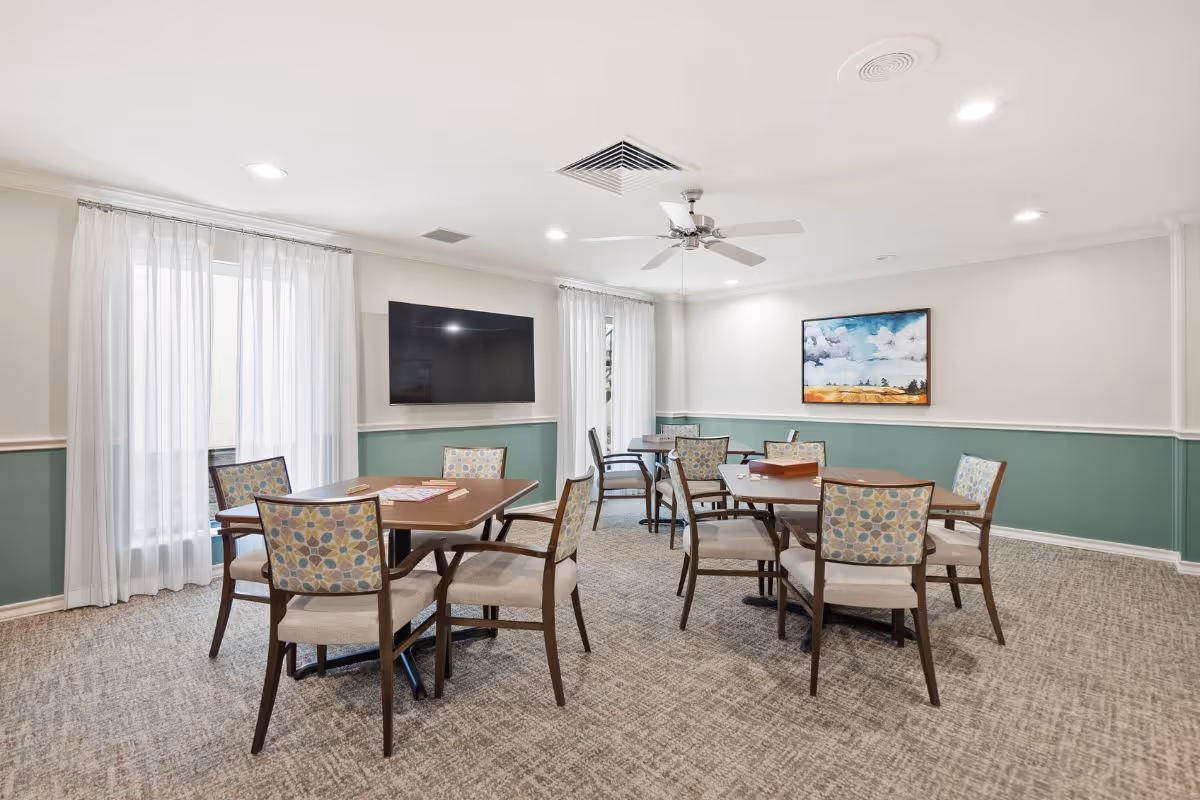 Bright communal dining/activity room with several tables and patterned chairs, a wall-mounted TV, and artwork on the wall.