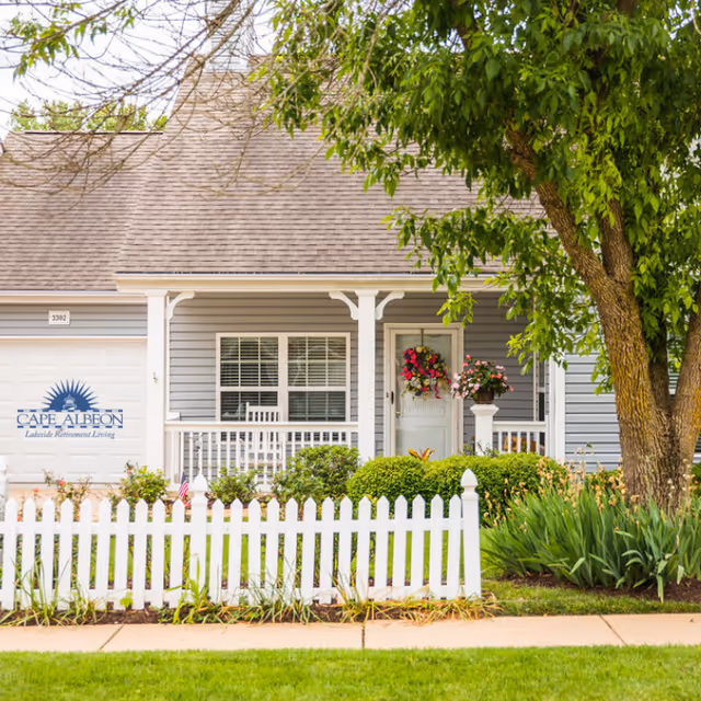 Front exterior view of a single-story house with gray siding, a white picket fence, a porch with white railings, a door decorated with a floral wreath, and a large tree on the right side.