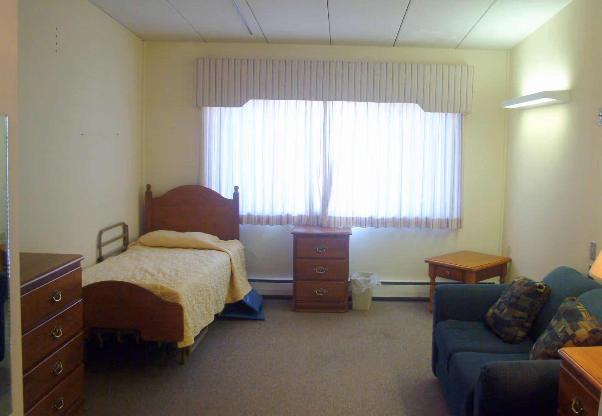 A simple, modestly furnished bedroom in a senior living facility with a single wooden bed covered with a beige quilt, a wooden dresser, a nightstand, a small table, a blue couch with patterned cushions, and a large window with white curtains letting in natural light.