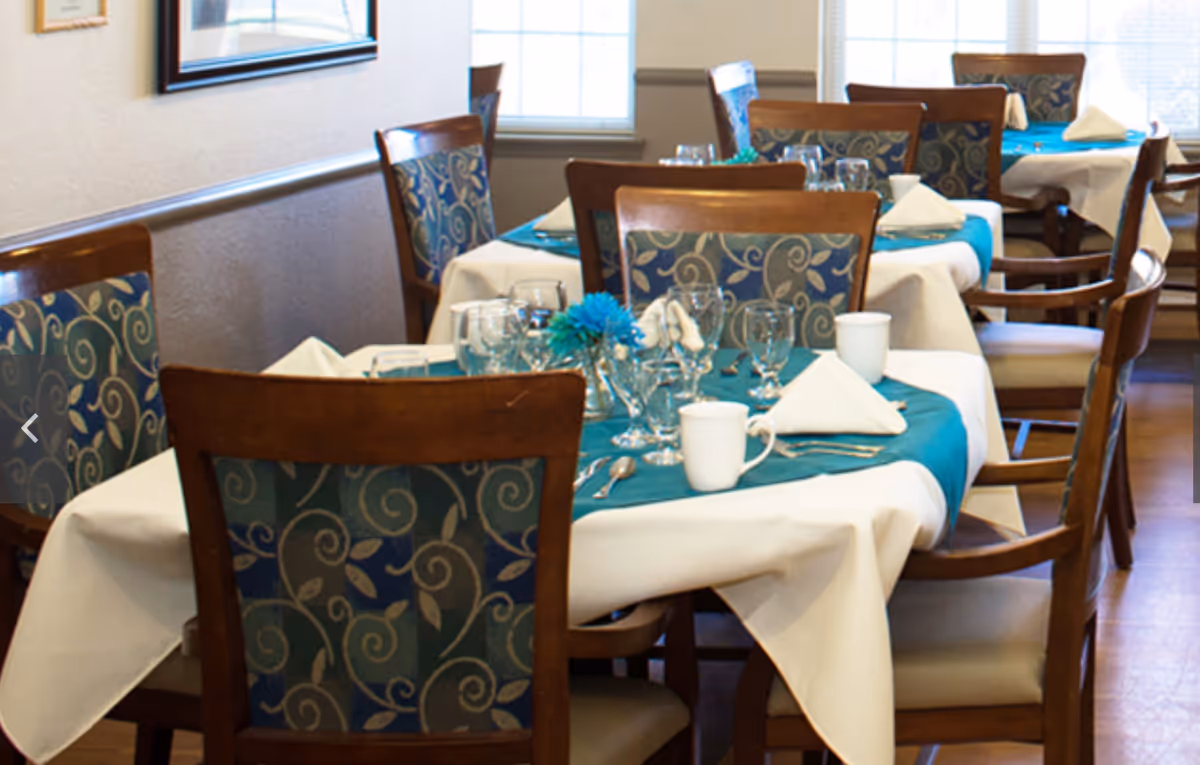 Dining room with tables set for a meal, featuring white tablecloths with teal runners, wooden chairs with patterned upholstery, glassware, white napkins folded in a triangle, and white mugs. The room has windows letting in natural light.