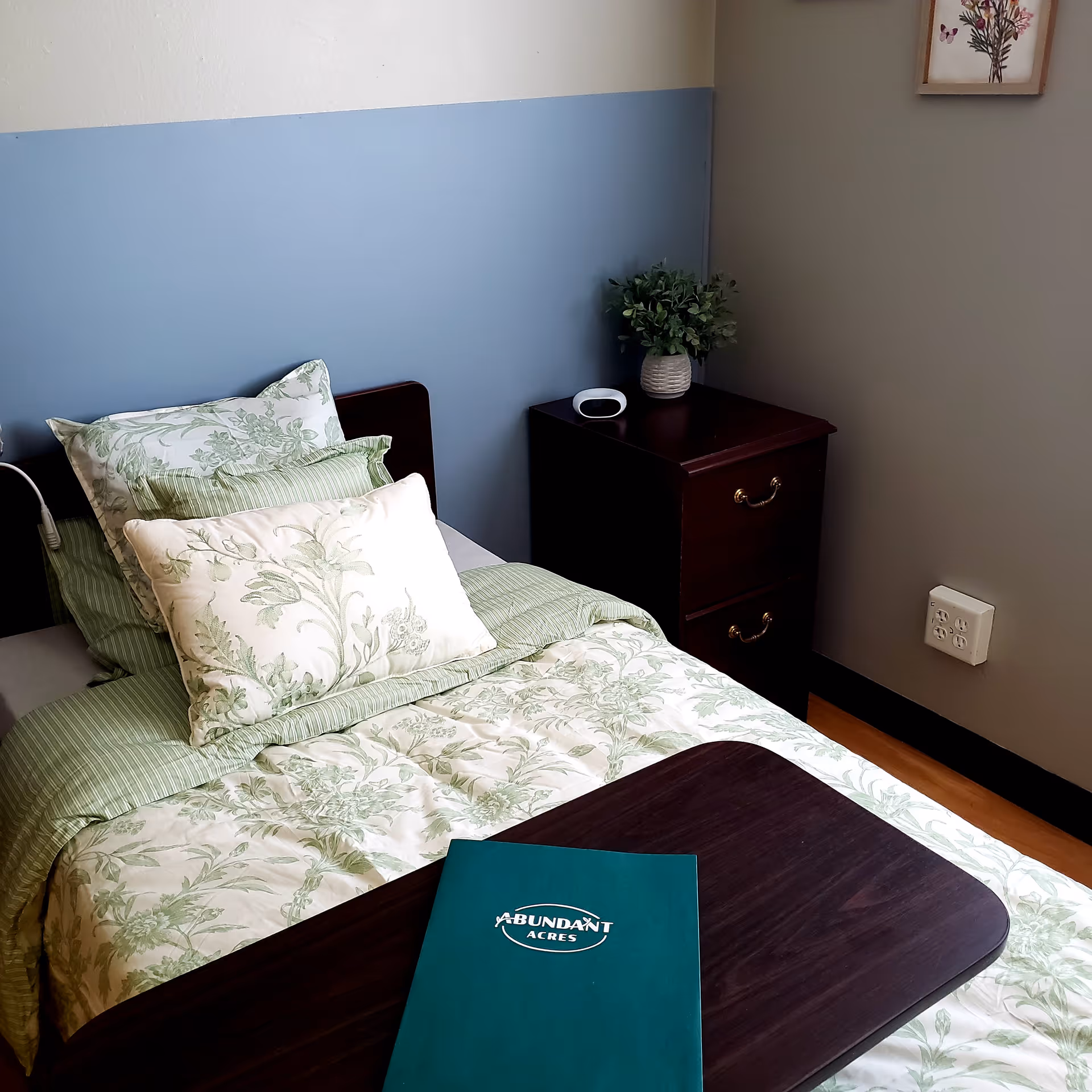 A neatly made single bed with green and white floral bedding and multiple pillows in a room with a blue and gray wall. Next to the bed is a dark wooden nightstand with a small potted plant and a white device on top. A dark wooden overbed table is positioned over the bed with a green folder labeled 'Abundant Acres'.