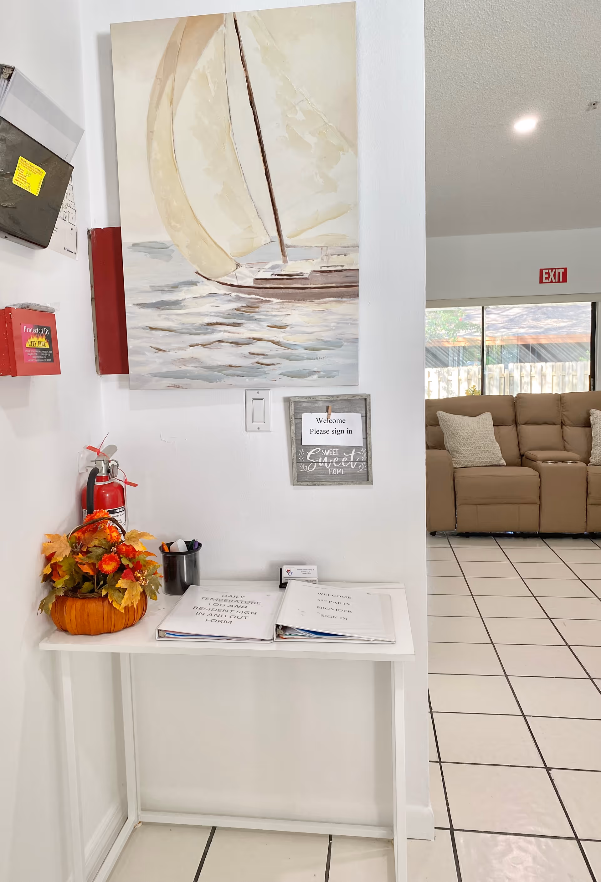 A small white table against a white wall with a fall-themed pumpkin decoration, a fire extinguisher, and open sign-in binders. Above the table is a painting of a sailboat on water and a framed sign that says 'Welcome Please sign in Sweet Home.' In the background, there is a beige sectional sofa with pillows and a large window with an exit sign above it.