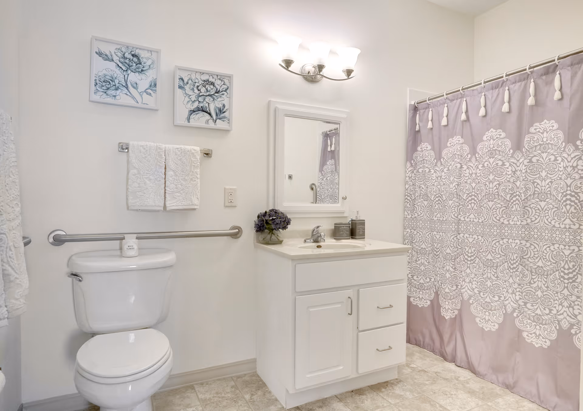 A clean and bright bathroom featuring a white toilet with a safety grab bar, a white vanity with a sink and mirror above it, a light fixture with three bulbs, two framed floral artworks on the wall, white towels hanging on a towel rack, and a purple shower curtain with a white intricate pattern.