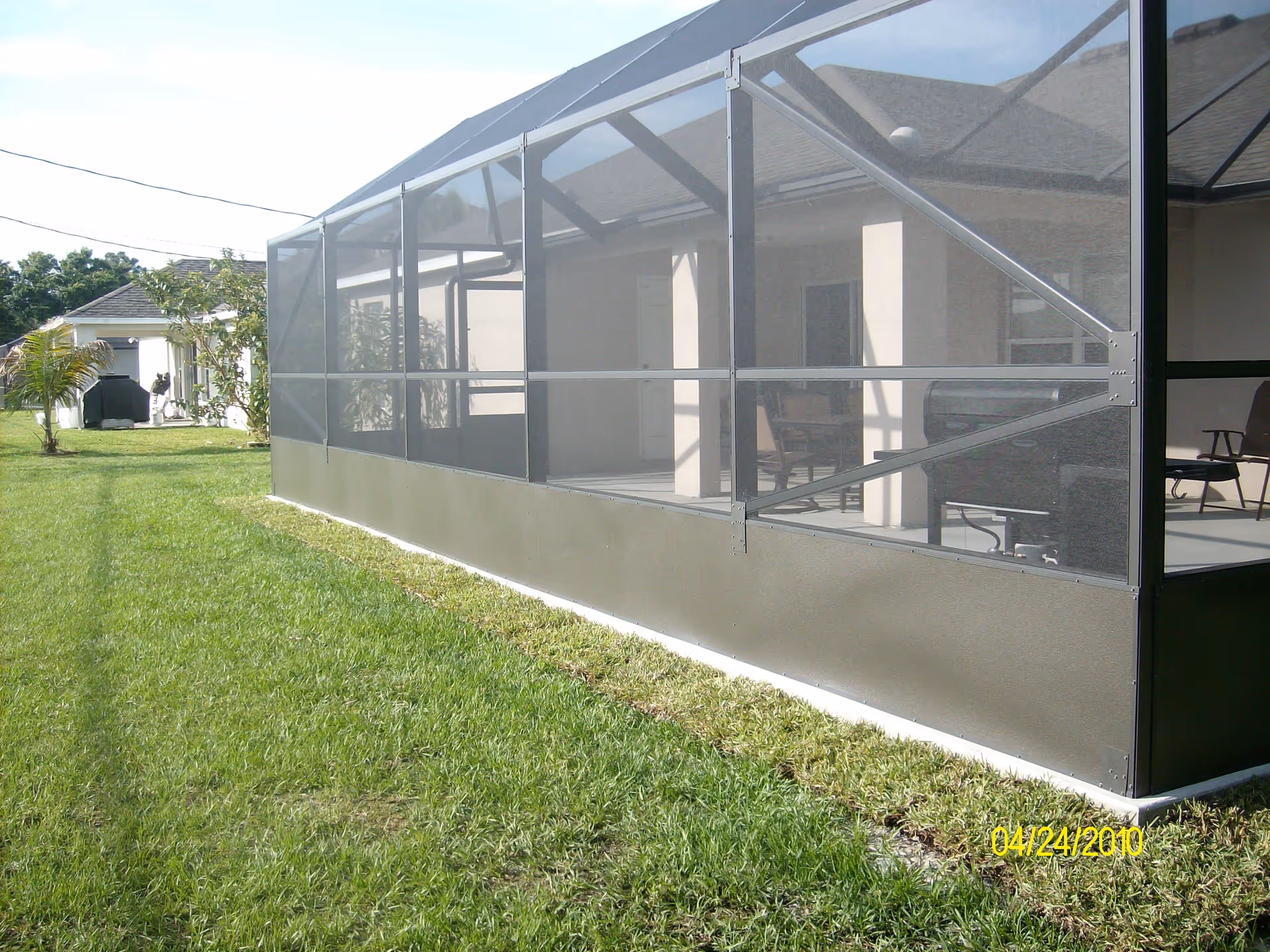 Outdoor view of a screened-in patio attached to a building, with green grass surrounding the area and some outdoor furniture visible inside the screened enclosure.
