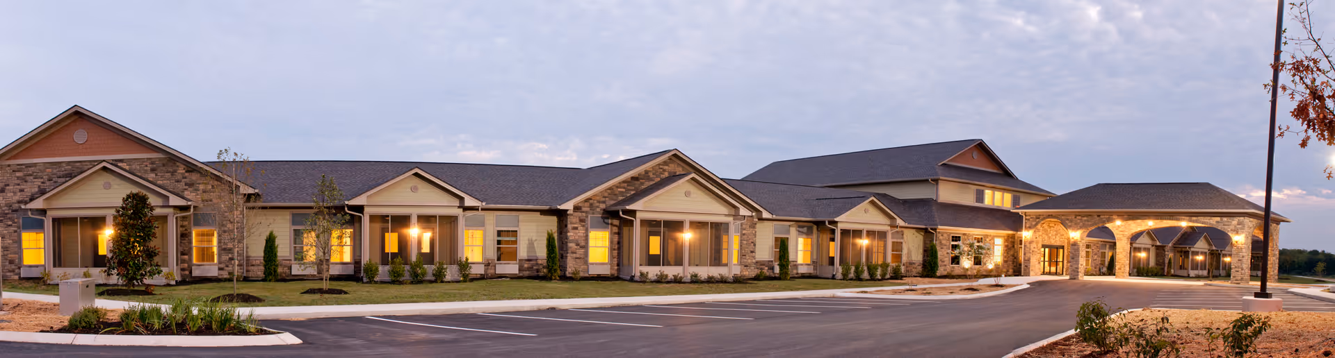 Wide exterior view of a single-story senior living facility building with stone and siding facade, multiple peaked roof sections, large windows with warm interior lighting, and a covered entrance with arches. The building is surrounded by a paved parking lot and landscaped areas with small trees and shrubs under a cloudy sky at dusk.